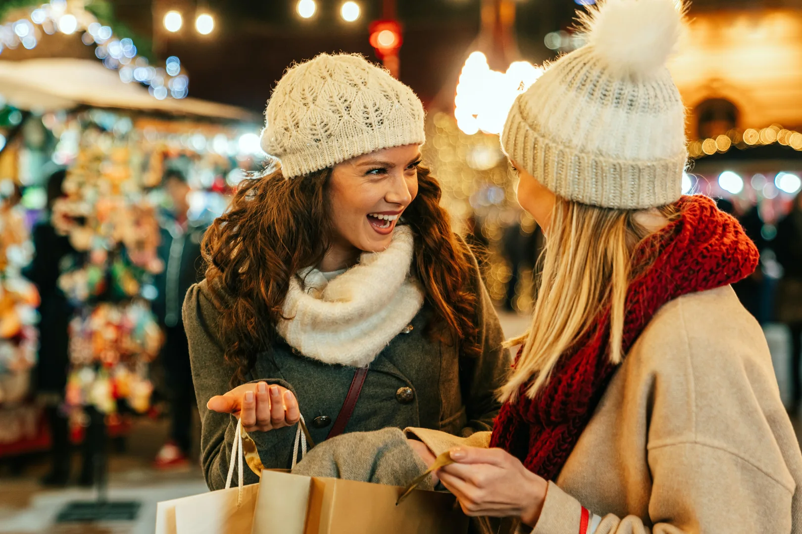 Two friends laughing together with shopping bags at a Christmas market under strings of lights